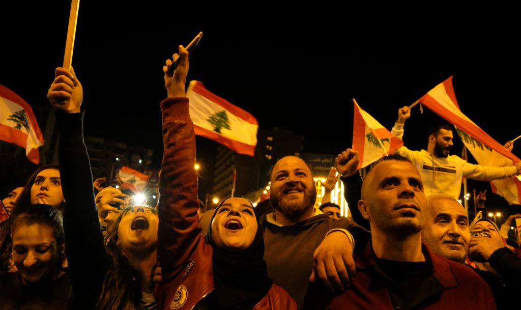 Mielenosoituksessa huutavia ja hymyileviä miehiä ja naisia, taustalla Libanonin punavalkoisia lippuja, joissa keskellä vihreä libanoninsetripuu. Ropande och leende män och kvinnor på en demonstration, i bakgrunden syns Libanons rödvita flaggor med den gröna libanoncedern i mitten.