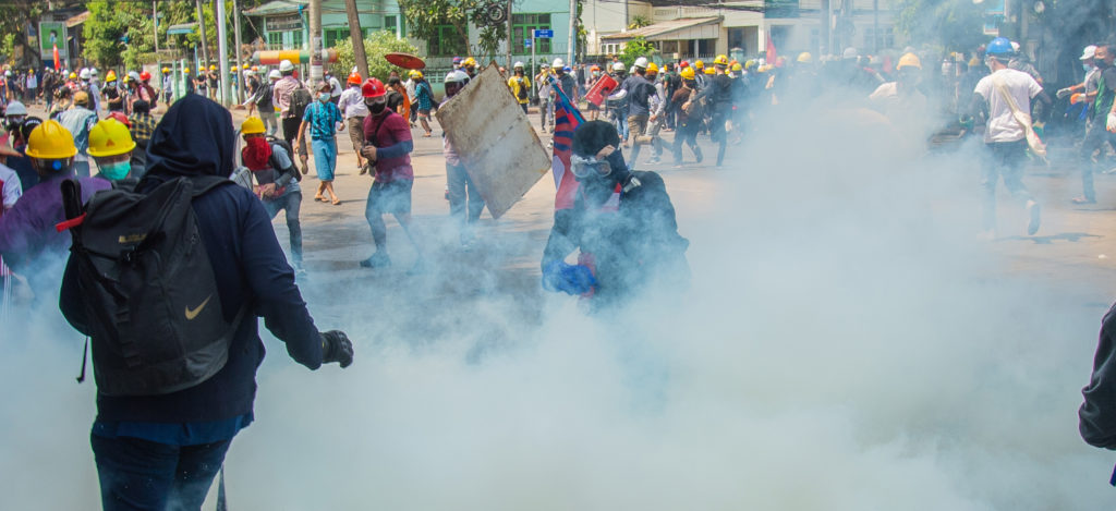 Mielenosoittajat juoksevat karkuun, etualalla poliisi kaasunaamarissa ja iso pilvi kyynelkaasua.