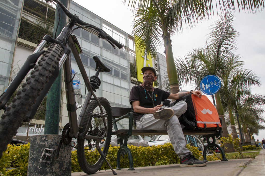 Mies istuu penkillä vieressään maastopyörä ja oranssi ruoankuljetusreppu.