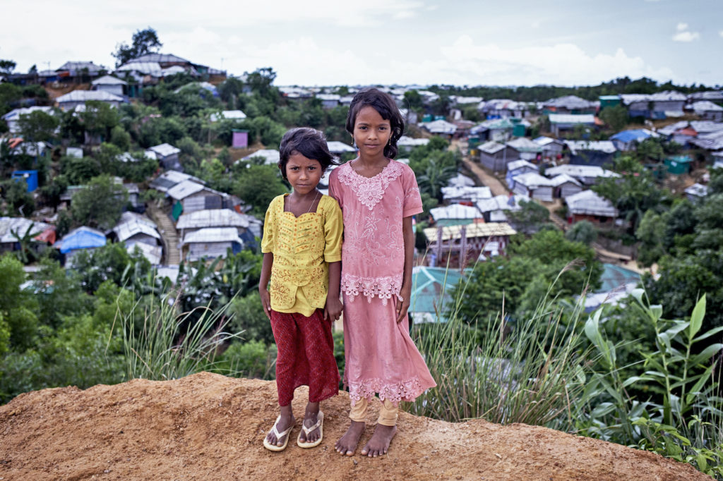 Kaksi tyttöä seisoo mäen päällä takanaan pakolaisleirin kattoja.