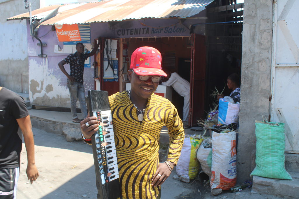 Singelilaulaja Mzee Rashidi Mbarak kävelee kadulla kosketinsoitin kainalossaan takanaan kadunvarsikauppoja.