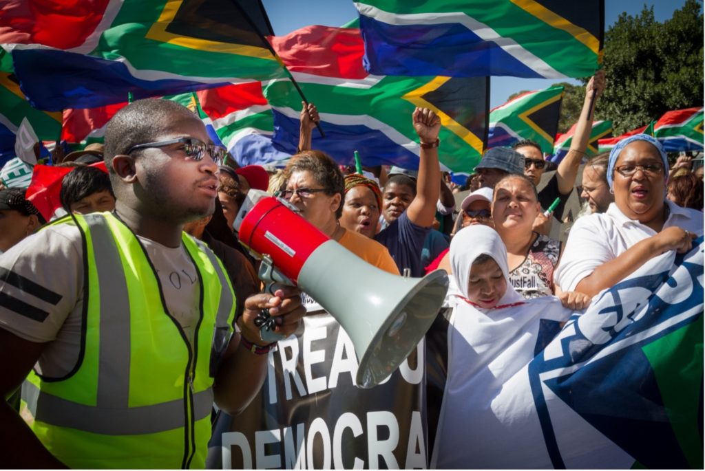 Ihmisiä Etelä-Afrikan lippujen kanssa mielenosoituksessa, etualalla henkilö megafonin kanssa.