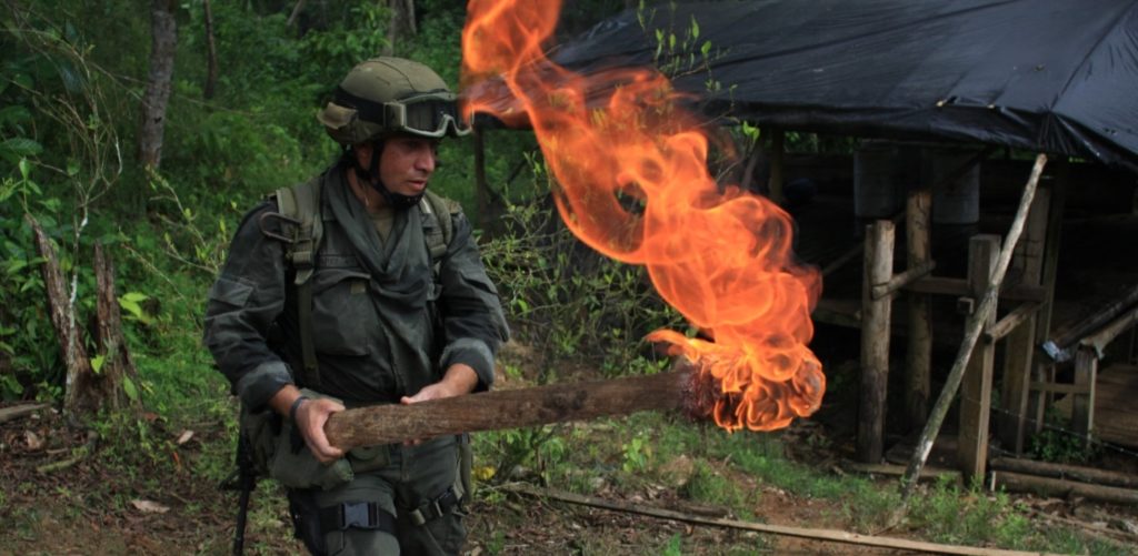 Sotilasasuinen mies pitää kädessään palavaa puuta.