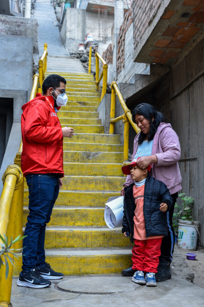 Maskiin ja punaiseen takkiin pukeutunut mies sekä äiti ja poika seisovat keltaisten portaiden edessä.