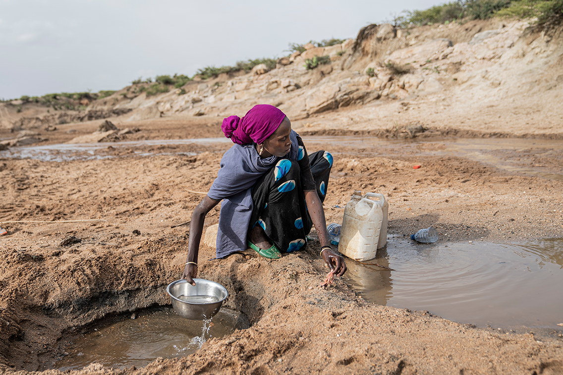 Laiha afrikkalainen nainen kauhoo kyykyssä vettä kahdesta maassa olevasta isosta lätäköstä.