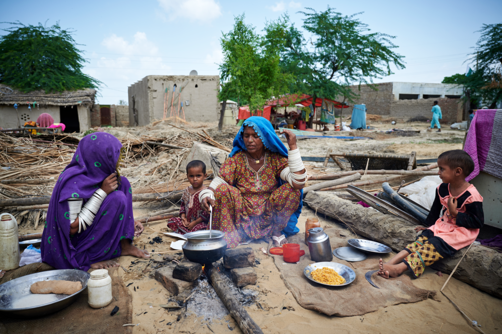 Kaksi naista ja kaksi lasta tekevät ruokaa maassa avotulella. Maassa on puunrunkoja ja taustalla vaurioituneita rakennuksia.