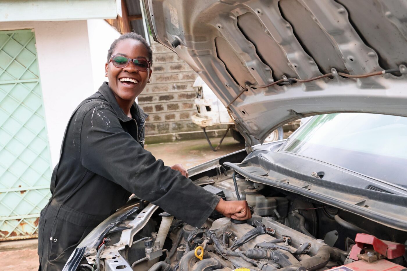 Nuori naurava nainen työtakissa, käsi auton konepellin alla olevissa osissa.
