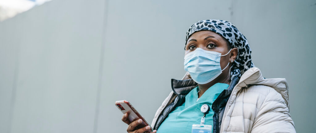 Tummaihoinen nainen terveydenhoitajan asussa kännykkä kädessään.