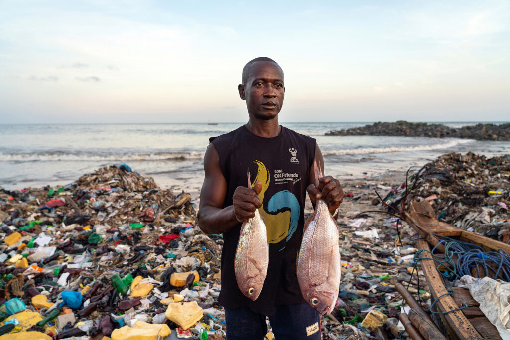 Mies esittelee kahta kalaa rannalla, joka on täynnä muovirojua.