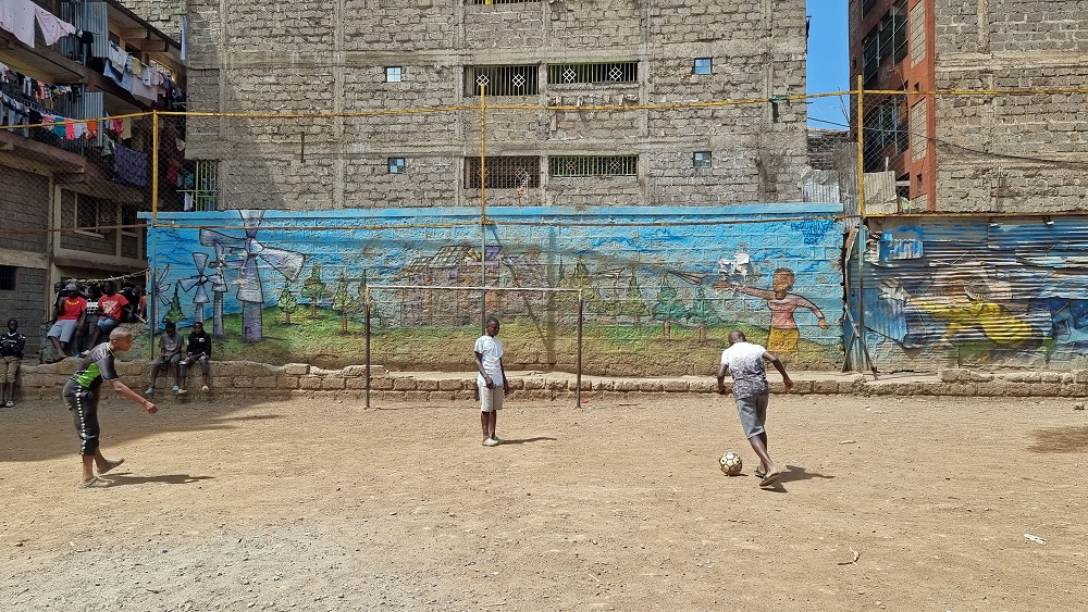 Kolme varhaisnuorta pelaa jalkapalloa, talon seinään on maalattu muraali, joka näkyy tuulimyllyjä.