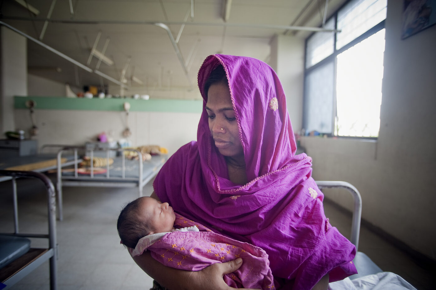 Pinkkiin huiviin ja mekkoon pukeutunut nainen pitää vauvaa sylissään klinikalla taustallaan rautasänkyjä.