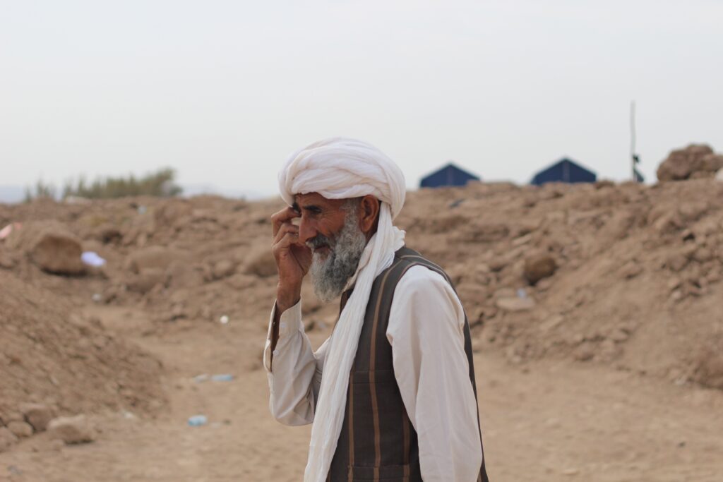 Ikääntynyt mies jolla on valkea turbaani ja paita sekä ruskea liivi seisoo sivuttain, taustalla hiekkaa.