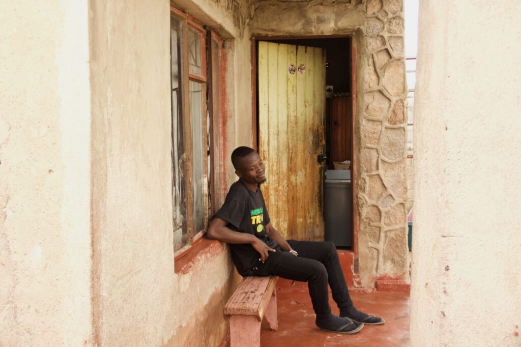Simbarashi Nyamasi istuu kotitalonsa edustalla sivuttain kameraan selkä ikkunaa päin, kuvan taustalla ovi, joka johtaa sisälle.