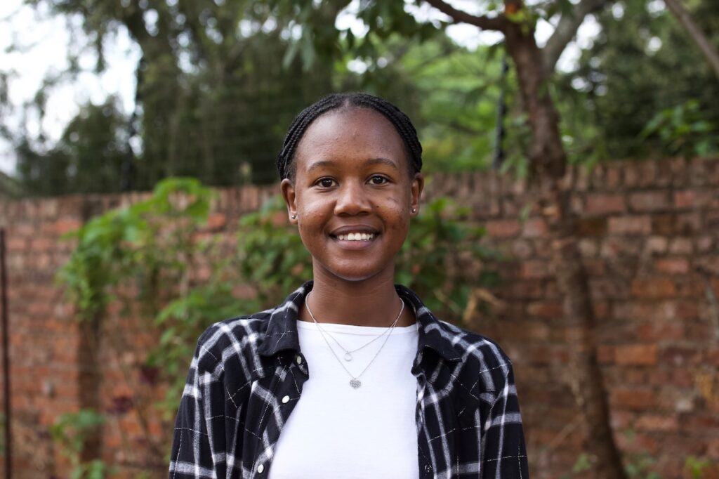 Nyasha Ngandu hymyilee kameralle taustanaan punatiilimuuri ja puita.