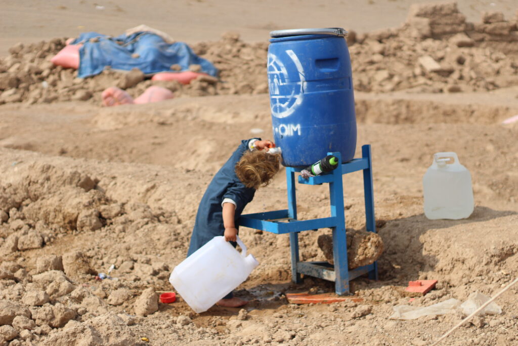 Lapsi valkoisen vesikanisterin kanssa sinisen vesisäiliön edessä, taustalla hiekkaa ja jätettä.