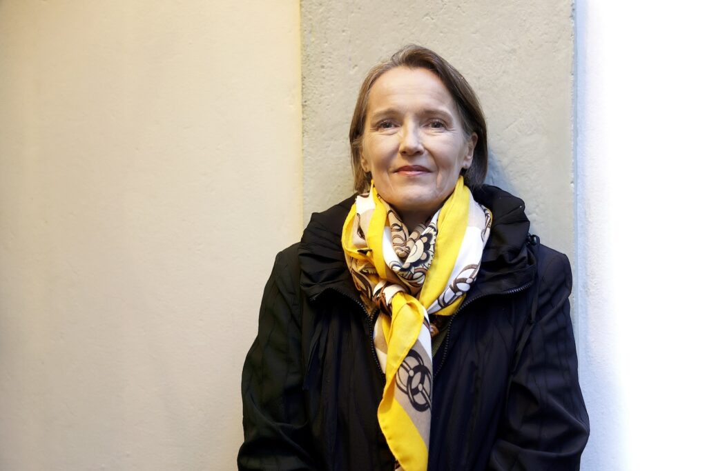 Liisa Laakso seisoo vaaleaa seinää vasten ja katsoo kameraan kaulassa iso kelta-ruskea-valkoinen kaulaliina.