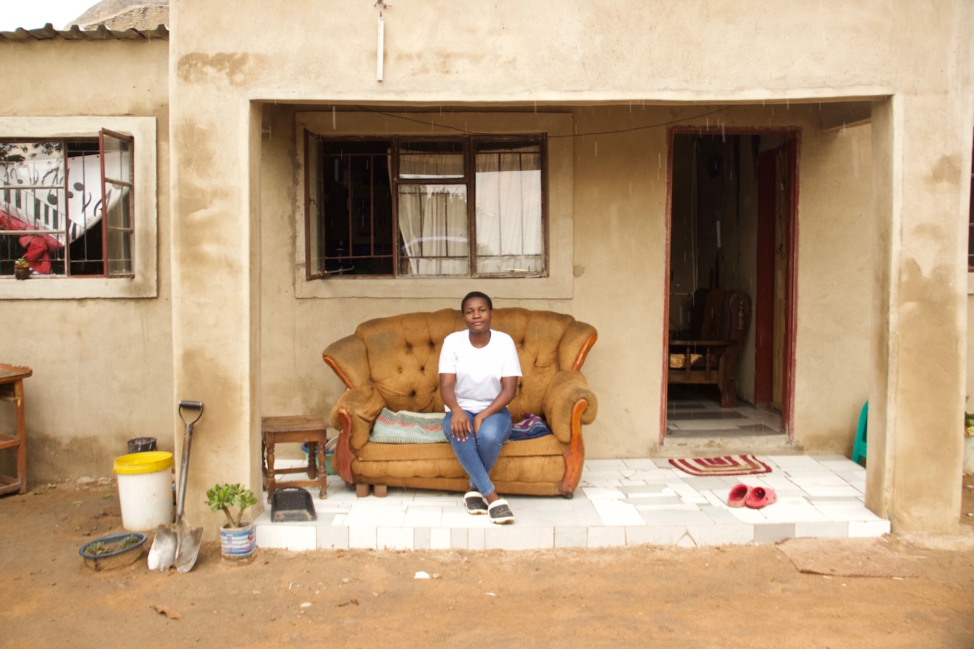 Faith Mawanda istuu nahkasohvalla kotitalonsa ulko-oven edustalla takaan ristikoitu ikkuna, edessä maassa muun muassa lapio, kukkaruukku ja muoviroskis.