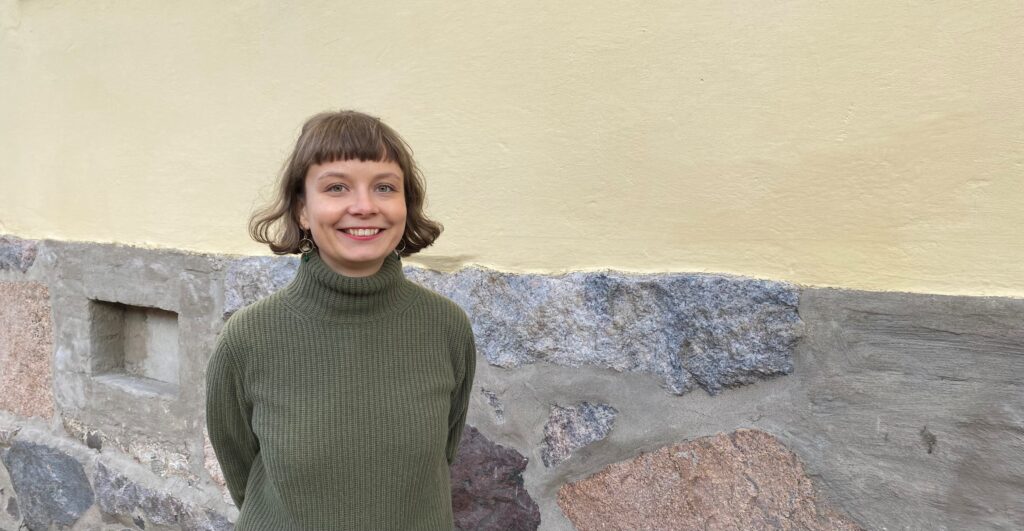 Ruskeahiuksinen nuori nainen vihreänharmaassa villapaidassa seisoo, nainen hymyilee, taustalla kivinen rakennus.