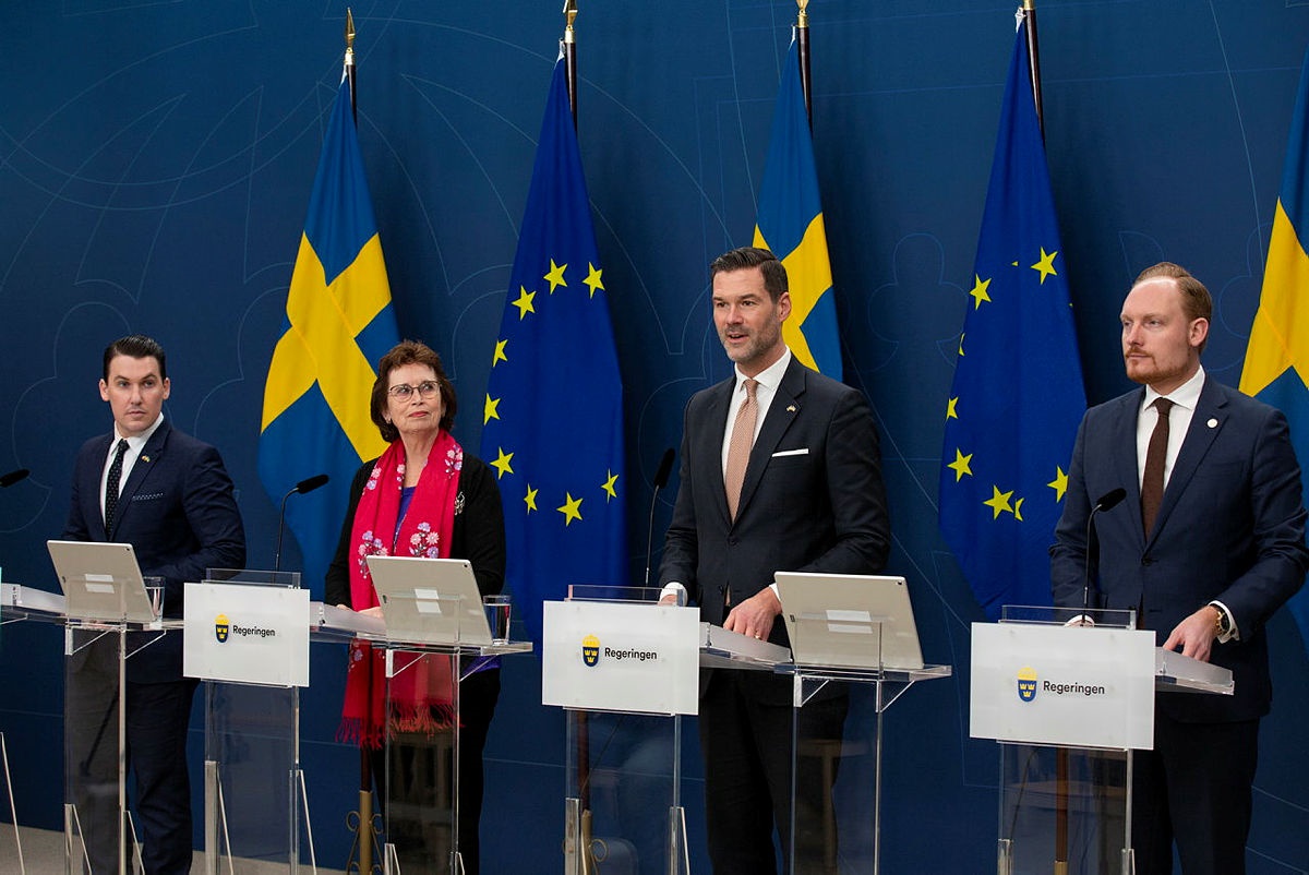 kolme miestä tummissa puvuissa ja yksi nainen punaisessa puserossa seisoo rinnakkain, taustalla Ruotsin ja EU:n liput.