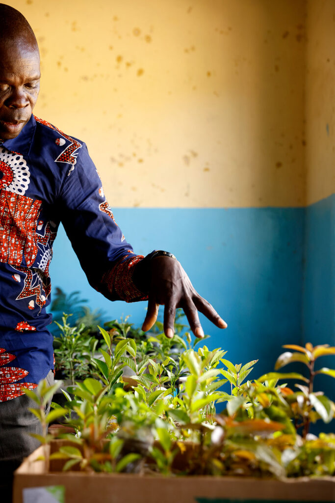 Puolikas kuva värikkääseen paitaan pukeutuneesta miehestä, joka osoittaa vasemmalla kädellään kasvitaimia.