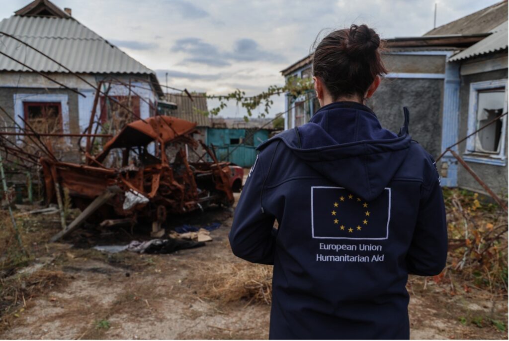 Nainen seisoo selin kameraan päällään Euroopan unionin tunnuksin varustettu huppari päällään edessään vaurioituneita taloja ja palanut auto.